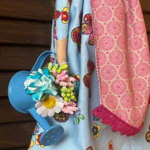 May include: A blue watering can with a handle and a spout, decorated with colorful flowers and green leaves. The watering can is hanging from a blue and white fabric with a butterfly pattern.