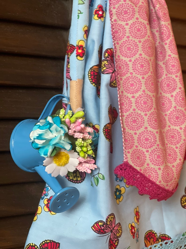 May include: A blue watering can with a handle and a spout, decorated with colorful flowers and green leaves. The watering can is hanging from a blue and white fabric with a butterfly pattern.