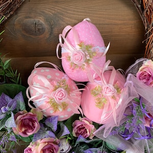 Op de afbeelding: Drie roze stoffen paaseieren met witte stippen en roze lintaccenten. Elk ei heeft een roze roos en groene bladeren. De eieren zijn gerangschikt op een houten oppervlak met paarse en groene bloemen.