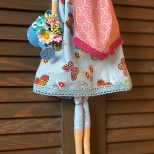 May include: A handmade doll with a blue dress with butterflies and flowers, a pink scarf, and blue polka dot legs. The doll is holding a blue watering can with flowers.