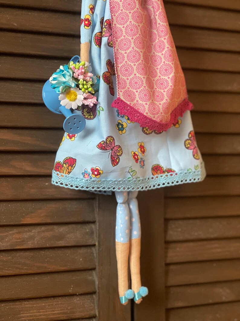 May include: A handmade doll with a blue dress with butterflies and flowers, a pink scarf, and blue polka dot legs. The doll is holding a blue watering can with flowers.
