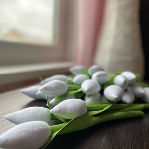 May include: A bunch of white fabric tulips with green fabric stems and leaves. The tulips are arranged on a dark brown wooden surface. The background includes a window and blurred objects.