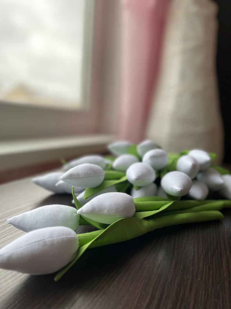 May include: A bunch of white fabric tulips with green fabric stems and leaves. The tulips are arranged on a dark brown wooden surface. The background includes a window and blurred objects.