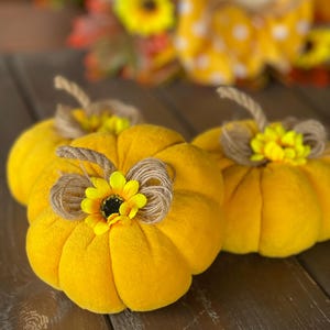 Pode incluir: Três abóboras de pelúcia amarelas com caules de barbante marrom e detalhes de girassol amarelo. As abóboras são dispostas em uma superfície de madeira, criando uma exibição decorativa com tema de outono.