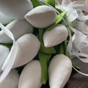 May include: A close-up of a bouquet of white fabric tulips with green fabric leaves and white ribbon. The tulips are tightly clustered, with the white petals contrasting against the green leaves and white ribbon. The bouquet is likely handmade.