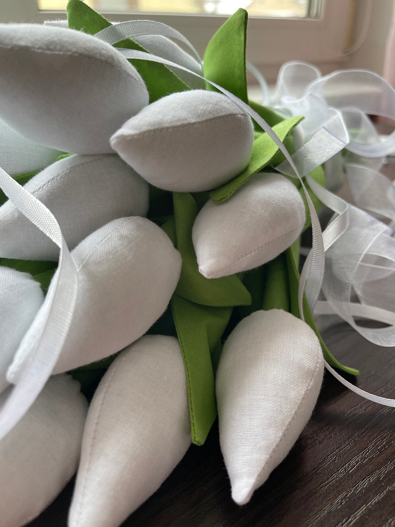 May include: A close-up of a bouquet of white fabric tulips with green fabric leaves and white ribbon. The tulips are tightly clustered, with the white petals contrasting against the green leaves and white ribbon. The bouquet is likely handmade.