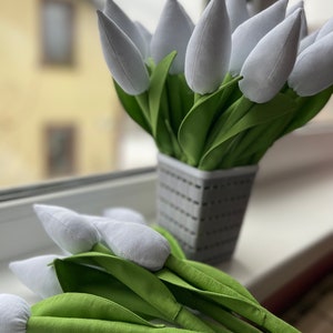 May include: A display of white fabric tulips with green fabric leaves, presented in a grey woven basket-style pot. Some tulips are placed on a windowsill. The background shows a blurred view of a building.