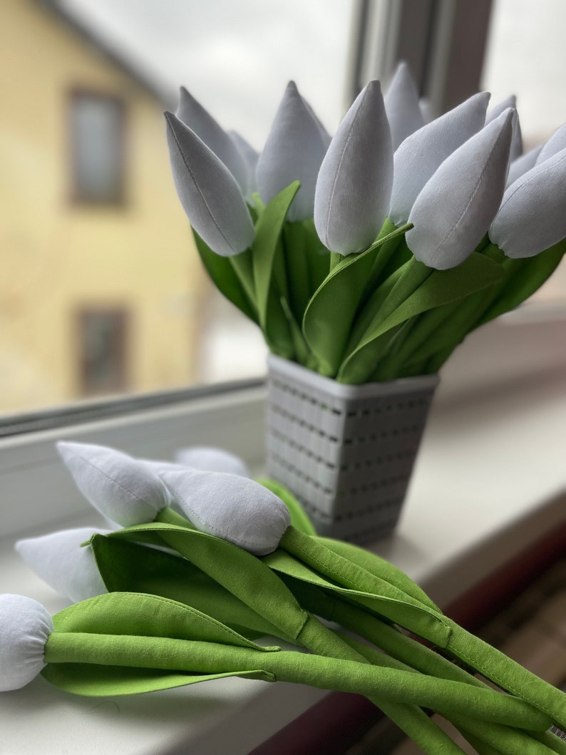 May include: A display of white fabric tulips with green fabric leaves, presented in a grey woven basket-style pot. Some tulips are placed on a windowsill. The background shows a blurred view of a building.