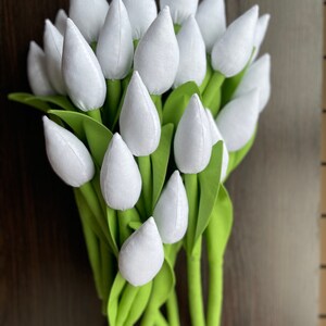 May include: A bunch of white fabric tulips with green stems and leaves. The tulip heads are a soft white, and the leaves are a bright green. The stems are a darker green, set against a dark brown background.