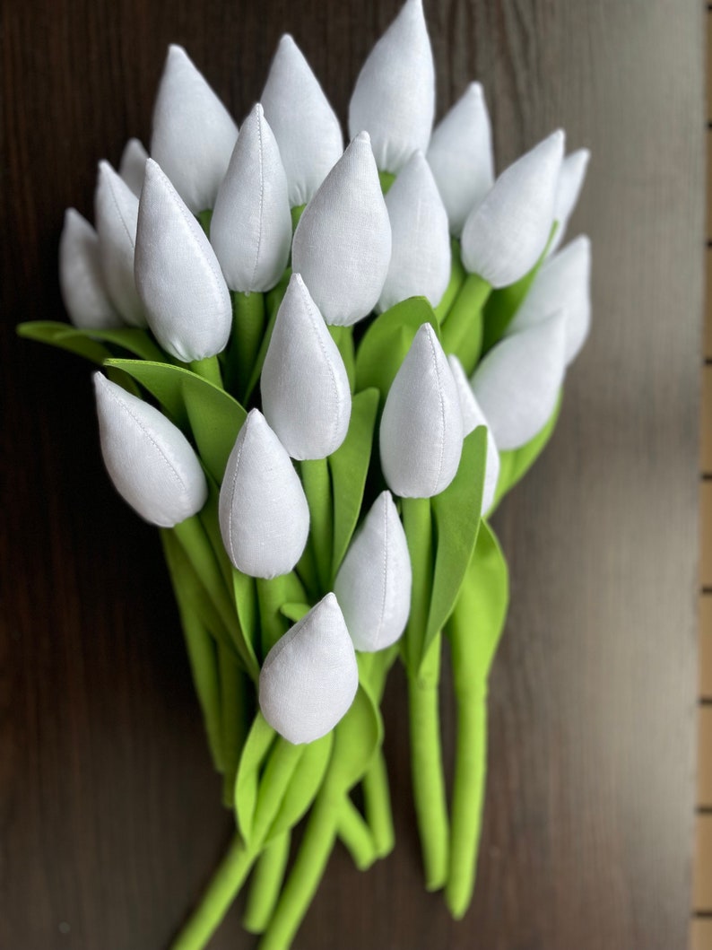 May include: A bunch of white fabric tulips with green stems and leaves. The tulip heads are a soft white, and the leaves are a bright green. The stems are a darker green, set against a dark brown background.