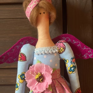 May include: A handmade doll with a blue dress, pink flower, and wings. The doll has brown hair tied with a pink ribbon and a white polka dot dress.