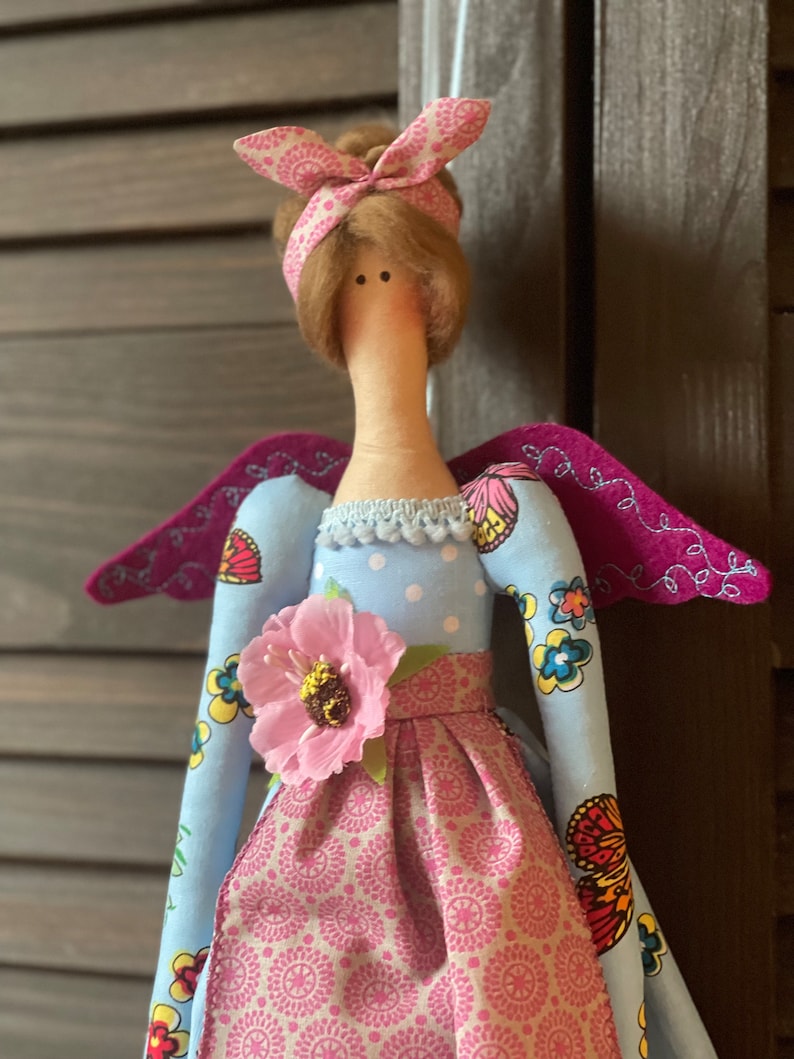 May include: A handmade doll with a blue dress, pink skirt, and wings. The doll has brown hair and a pink headband. The doll is wearing a flower on her dress.