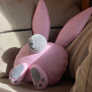 May include: A pink bunny-shaped pillow with white polka dots and gray felt feet and ears. The pillow is lying on a brown couch.