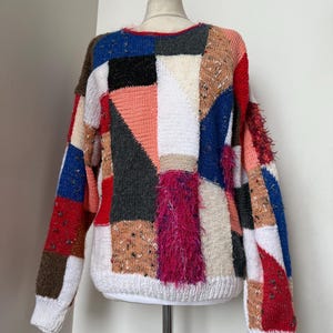 May include: A colourful patchwork sweater with a variety of textures. The sweater features squares of red, blue, pink, white, and brown. The sleeves and hem are white, and the sweater is displayed on a mannequin.