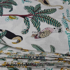 Op de afbeelding: Witte stof met een herhaald patroon van toekans, luipaarden en palmbomen in groen, geel en bruin. De stof is gevouwen en toont meerdere lagen van het gedrukte ontwerp. Het algemene thema is tropisch.