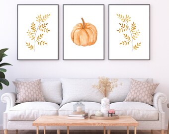 May include: Three framed watercolor prints featuring a single orange pumpkin and two branches of gold leaves. The prints are displayed on a white wall above a white sofa with two pillows.