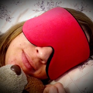 Op de afbeelding: Een close-up van een persoon die slaapt met een roze slaapmasker over hun ogen. Het masker heeft een zwarte rand en is gemaakt van zacht, pluche materiaal.