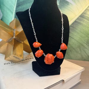 May include: A silver chain necklace featuring large, irregularly shaped, orange-red coral stones. The necklace is displayed on a black jewelry bust against a backdrop of green and white foliage.