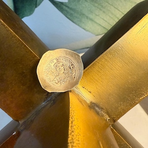 May include: A small, silver-colored coin with an embossed design rests on a gold-colored, star-shaped decorative object. The coin's design includes text and a central emblem. The gold object has a textured surface and is positioned against a backdrop of green and white.