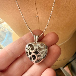 Puede incluir: Un collar con colgante en forma de corazón plateado con un diseño calado abierto. El colgante cuelga de una delicada cadena de bolas. El corazón tiene una apariencia tridimensional y se sostiene en una mano.