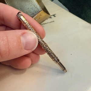 Könnte beinhalten: Ein antiker Bleistift mit einem detaillierten floralen Muster. Der Bleistift besteht aus goldfarbenem Metall und hat oben eine kleine Schlaufe. Die Spitze des Bleistifts ist silberfarben. Der Bleistift wird in einer Hand gehalten.