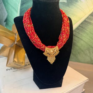 Puede incluir: Un collar de cuentas rojas con detalles dorados y un colgante dorado. El collar se exhibe sobre un busto negro. El colgante tiene detalles intrincados y una borla colgante.