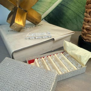May include: A set of clear crystal glass knife rests in a decorative box. The box has a black and white patterned exterior and a red interior. The knife rests are arranged on a cream-colored fabric.