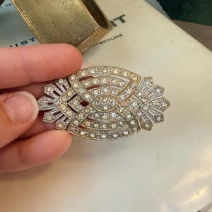 Op de afbeelding: Een zilverkleurige Art Deco broche met een geometrisch ontwerp, bezet met talrijke heldere strass-steentjes. De broche heeft een ovale vorm met symmetrische, waaiervormige verlengingen aan elk uiteinde. Het ontwerp is ingewikkeld en gedetailleerd.