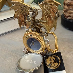 Puede incluir: Una figura de dragón dorada con las alas extendidas, sosteniendo un reloj de bolsillo con una esfera azul oscuro. El reloj está sujeto a una cadena dorada. Un pequeño compacto plateado con la inscripción "Mila Curli" está abierto.