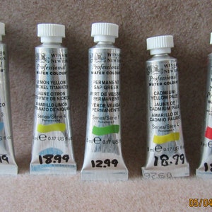 Peut inclure: Cinq tubes de peinture à l'eau Winsor & Newton Professional. Les tubes sont argentés avec des bouchons blancs et contiennent les couleurs suivantes : Bleu céruléen, Jaune citron, Vert sapin permanent, Jaune de cadmium pâle et Rouge de cadmium. Chaque tube est étiqueté avec le nom de la couleur, le numéro de série et le volume en millilitres.