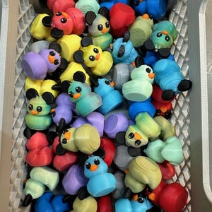 May include: A collection of colorful, 3D-printed duck figurines in a gray plastic bin. The ducks are various colors, including red, yellow, blue, green, and purple, with black ears and beaks. The bin is filled with the duck figurines.