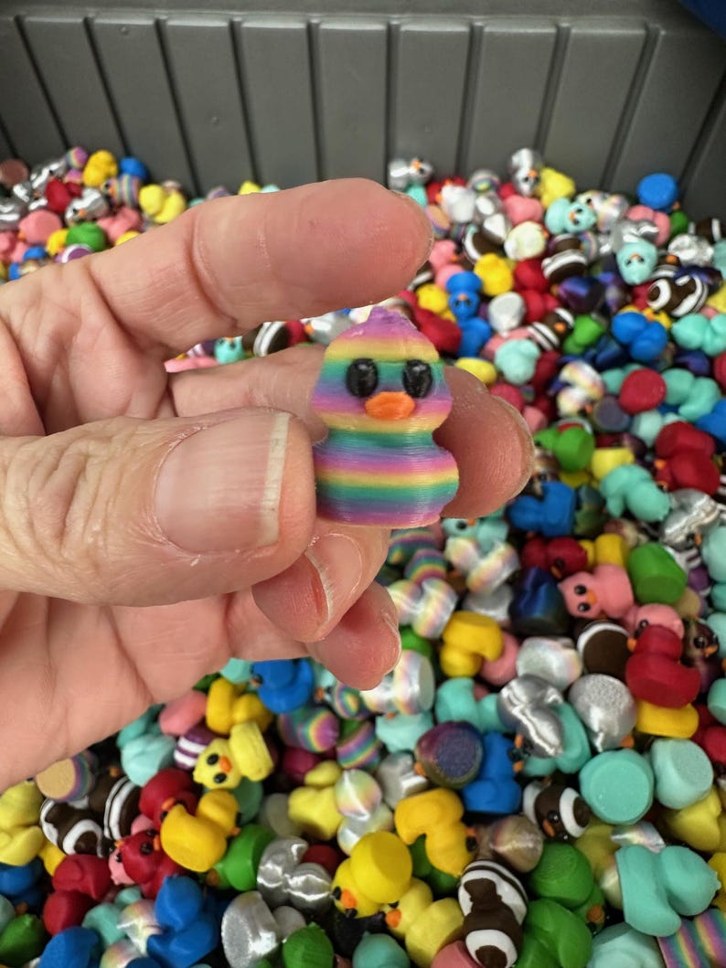 May include: A small, colorful duck figurine held in a hand, with a rainbow striped body and a purple head. The duck has black eyes and an orange beak. The background is filled with many other duck figurines in various colors.