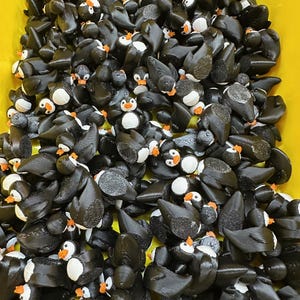May include: A large collection of small, black penguin figurines with white bellies, orange beaks, and white eyes. The penguins are densely packed together, filling a yellow container. The figurines appear to be 3D-printed.