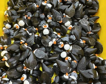 May include: A large collection of small, black penguin figurines with white bellies, orange beaks, and white eyes. The penguins are densely packed together, filling a yellow container. The figurines appear to be 3D-printed.
