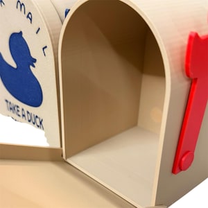 May include: A beige mailbox with a red flag and a blue duck graphic. The mailbox has the words "MAIL" and "TAKE A DUCK" printed on it. The mailbox is made of a durable material and has a spacious interior.