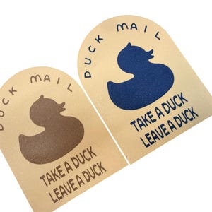 May include: Two beige, arched-top signs with the words "DUCK MAIL" and a duck silhouette. One sign has a brown duck, the other a blue duck. The text "TAKE A DUCK LEAVE A DUCK" is below the duck image.
