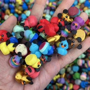 May include: A hand holds a collection of colorful, miniature mouse figurines. The mice are in various colors, including red, yellow, blue, and purple, with black ears and eyes. The background is filled with more of the same figurines.