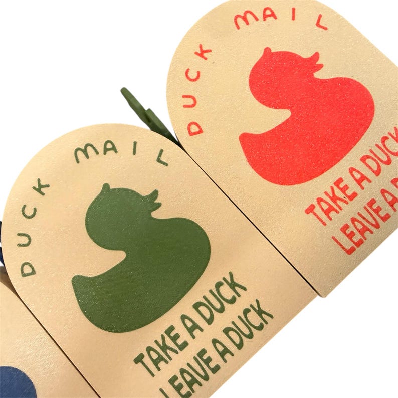 May include: Close-up of three beige, arched-top cards with a duck silhouette and the words "DUCK MAIL" and "TAKE A DUCK LEAVE A DUCK" printed in red and green. The cards have a textured appearance.