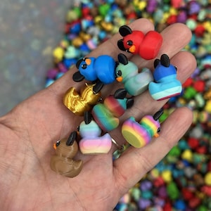 May include: A hand holds a collection of small, colorful duck figurines. The ducks are various colors, including red, blue, green, brown, gold, and rainbow patterns. Each duck has black ears and eyes. The background is a blur of other colorful objects.