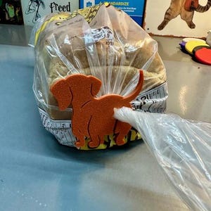 May include: A loaf of bread in a clear plastic bag, secured with an orange dachshund-shaped bag clip. The bag clip is made of a solid material. The background includes a sign that says "Our House Elf was Freed."