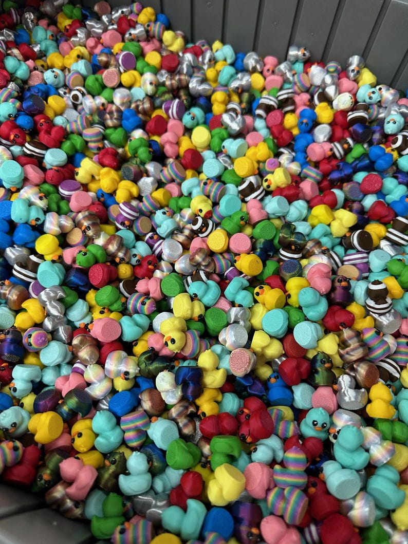 May include: A large assortment of colorful rubber duck toys. The ducks are in various colors including yellow, blue, red, green, and pink. Some ducks have striped patterns, while others have solid colors. The ducks are in a gray bin.