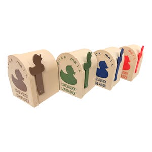 May include: A row of beige mailboxes with duck-shaped cutouts and handles. Each mailbox has a different colored duck and handle: brown, green, blue, and red. Text on the mailboxes reads "Duck Mail" and "Take a Duck, Leave a Duck."