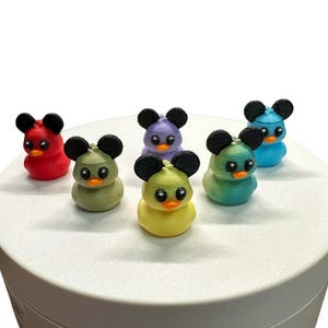 May include: A collection of six colorful duck figurines with mouse ears. The ducks are in various colors, including red, green, yellow, purple, and blue. Each duck has black mouse ears and orange beaks. The figurines are arranged on a white surface.