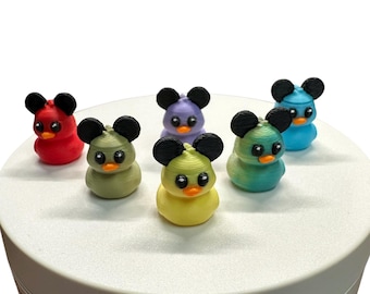 May include: A collection of six colorful duck figurines with mouse ears. The ducks are in various colors, including red, green, yellow, blue, and purple. Each duck has black ears and orange beaks. The figurines are arranged on a white surface.