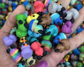 May include: A handful of colorful, miniature duck figurines in various colors, including green, blue, red, and purple. Each duck has a black beak and feet. The background is filled with more of the same figurines.
