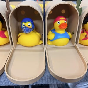 May include: Four yellow rubber ducks in decorative mailboxes. Each duck is dressed in a different costume: a pirate, a superhero, a lifeguard, and another pirate. The ducks have orange beaks and are inside beige mailboxes.