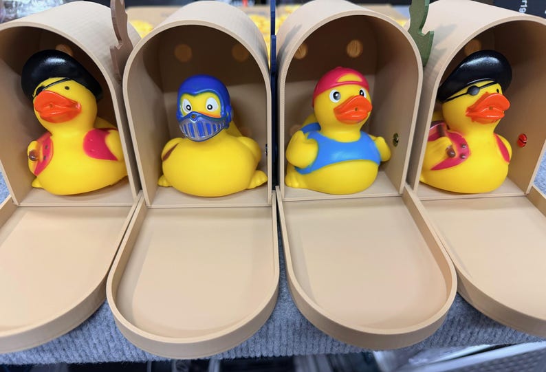 May include: Four yellow rubber ducks in decorative mailboxes. Each duck is dressed in a different costume: a pirate, a superhero, a lifeguard, and another pirate. The ducks have orange beaks and are inside beige mailboxes.