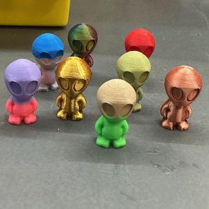 May include: A collection of small, colorful alien figurines. The figures are in various colors, including blue, green, red, gold, and purple. Each figurine has a rounded head with large oval eyes and a small body with arms and legs. The figurines are arranged on a gray surface.