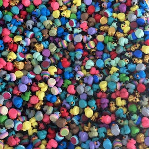 May include: A vibrant assortment of miniature duck figurines in various colors, including yellow, blue, red, and purple. The ducks have black eyes and some have additional decorative elements. The image is a close-up, showing the figurines densely packed together.