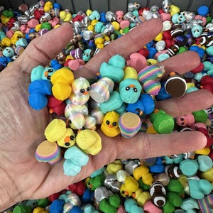 May include: A hand holding a collection of colorful miniature duck figurines. The ducks are various colors, including yellow, blue, green, and pink, with some featuring striped or patterned designs. The background is filled with more of the same figurines.